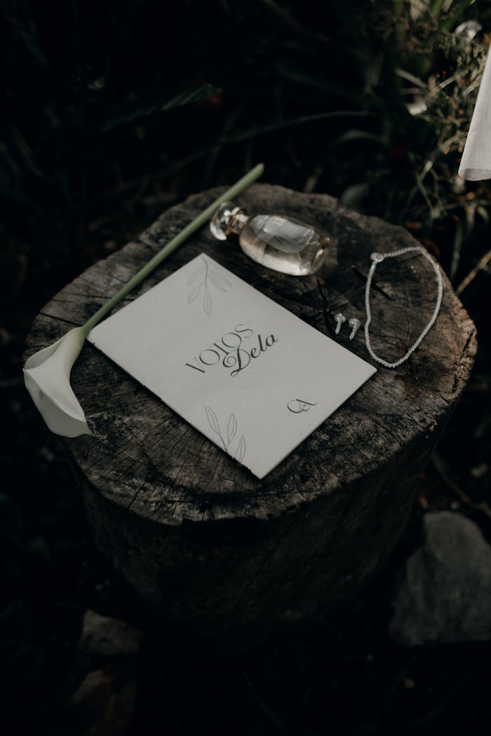 Stylish wedding invitation on tree stump with perfume, jewelry, and calla lily in natural setting.